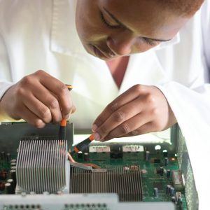 Concentrated African American technician wearing lab coat and conducting expertise of motherboard by using screwdrivers while working in service center