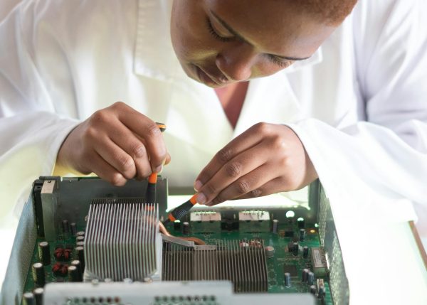 Concentrated African American technician wearing lab coat and conducting expertise of motherboard by using screwdrivers while working in service center